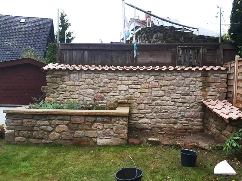 Eine Natursteinmauer mit Ziegeldachabdeckung steht in einem Garten. Vor der Mauer befindet sich ein kleines Beet, bepflanzt mit Kräutern und Blumen. Im Hintergrund sind Holzzäune und Häuser zu sehen.