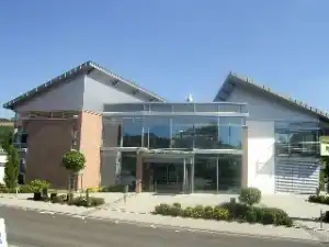 Ein modernes Gebäude mit viel Glas an der Vorderseite. Es besteht aus zwei verbundenen Gebäudeteilen mit schrägen Dächern. Vor dem Eingang wachsen kleine Bäume und Sträucher.