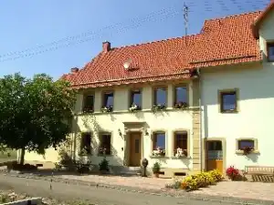 Ein langes, helles Wohnhaus mit rotem Ziegeldach. Vor dem Haus stehen Blumen und kleine Sträucher. Die Fenster sind mit Blumenkästen geschmückt.