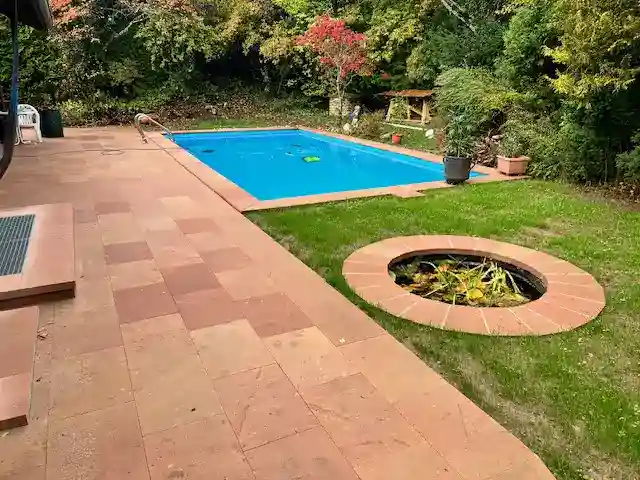 Ein rechteckiger, blauer Pool liegt in einem Garten. Daneben eine große Terrasse aus rötlichen Platten und ein kleiner, runder Teich mit Wasserpflanzen. Im Hintergrund dichte Bäume.