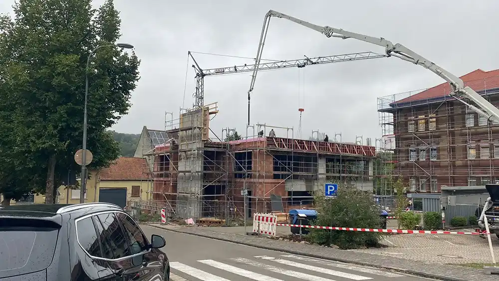 Eine Baustelle an einer Straße mit einem Gebäude im Rohbau und einem Baukran. Das Bauwerk ist von Gerüsten umgeben. Ein Auto parkt am Straßenrand, und Absperrbänder sichern die Baustelle.