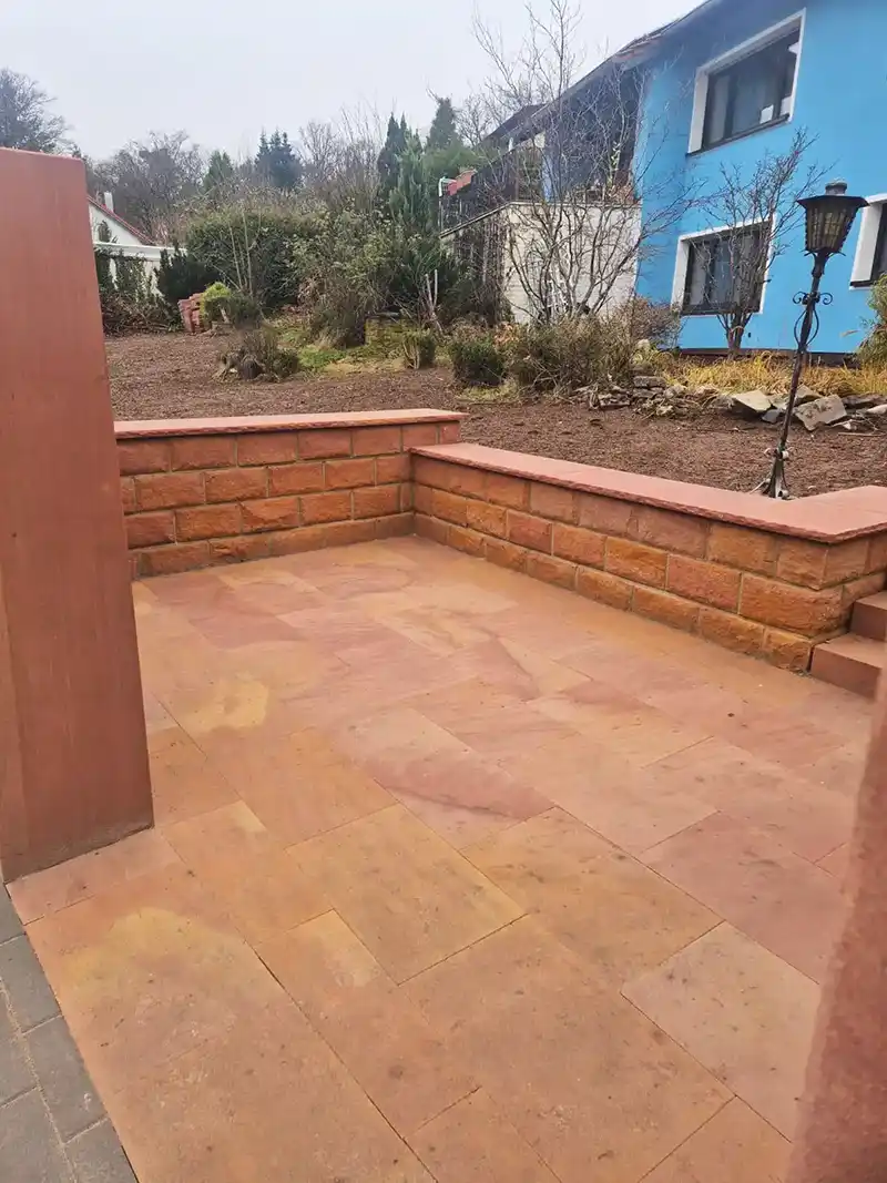 Eingangsbereich mit rötlichen Sandsteinplatten und niedrigen Steinmauern. Im Hintergrund ein Garten mit Sträuchern und ein blau gestrichenes Wohnhaus.