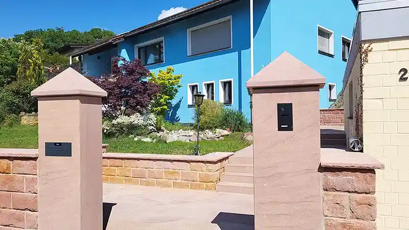 Hauseingang mit Sandsteinmauer, Torpfeilern und gepflegtem Vorgarten vor einem blauen Wohnhaus